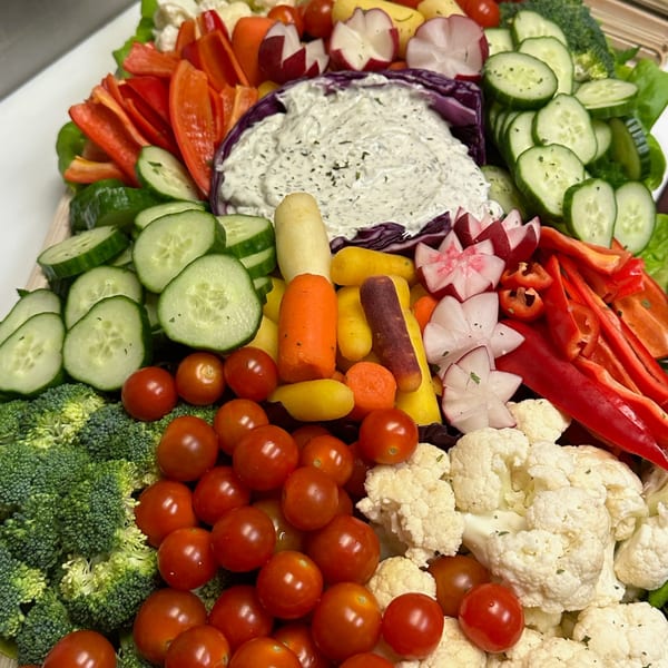 Colorful vegetable platter with broccoli, cherry tomatoes, cucumbers, bell peppers, radishes, and dip.